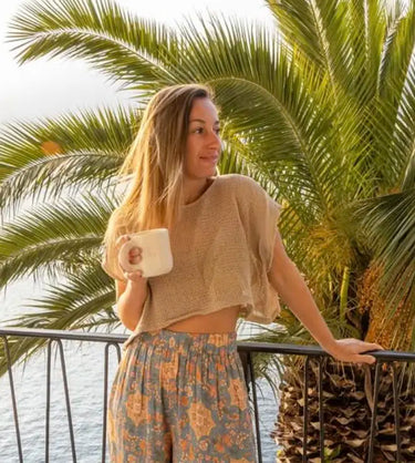 Customer Lucy enjoying morning coffee on balcony surrounded by palm trees, wearing relaxed natural clothing, representing Kiseau’s sustainable lifestyle