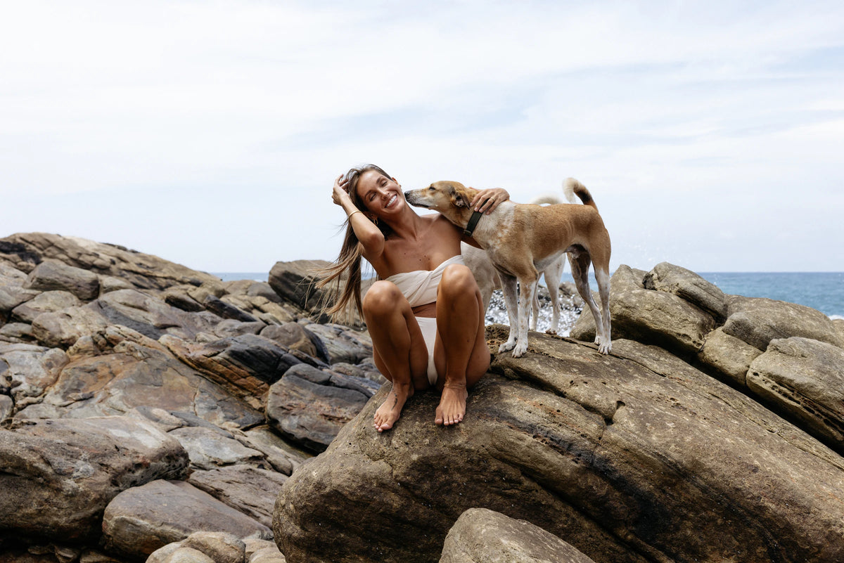 Model in Kiseau Natural Undyed Classic Thong, laughing as a dog nuzzles her, wide rock shelf beside the sea, seaweed-infused organic cotton