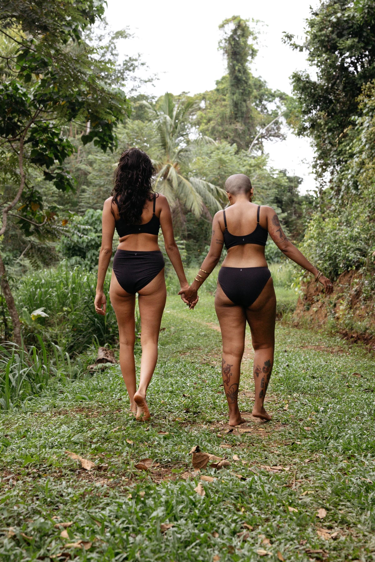 Two models in Kiseau Charcoal Everyday Bralettes, Cheeky Bikinis and High Waist Briefs, walking away, hands clasped, grassy jungle path