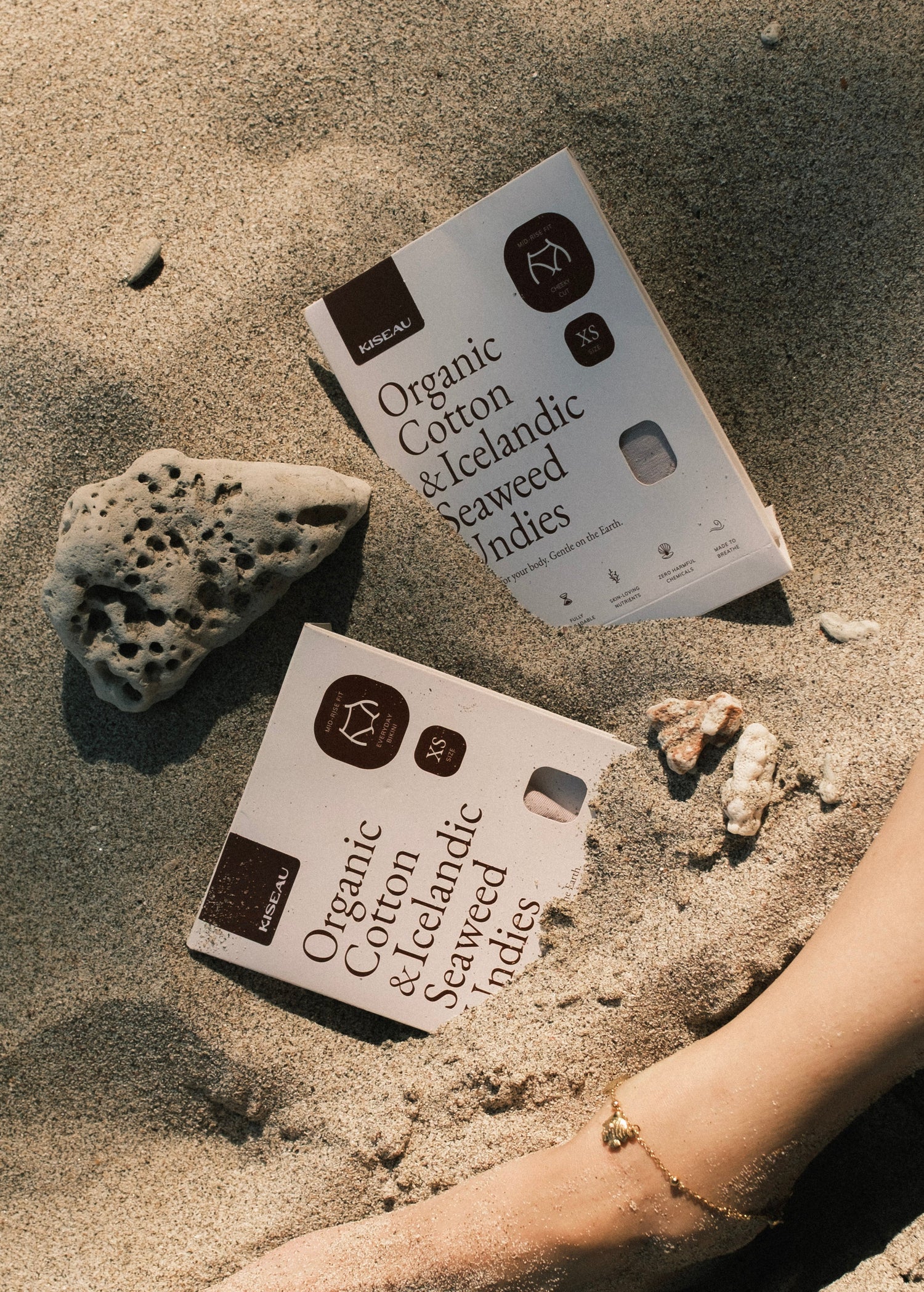Two boxes of Organic Cotton & Icelandic Seaweed Indies on sand with a hand reaching towards them.